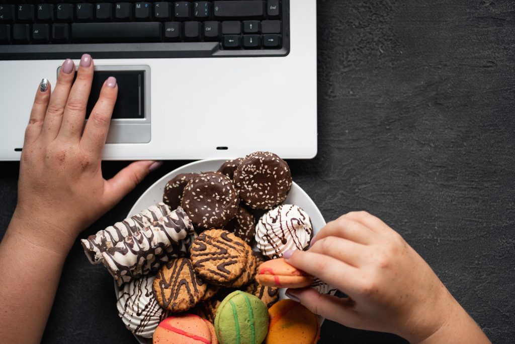 Woman eating cookies and coffee at workplace Unhealthy snack at work time. Compulsive indulgence, overeating, stress, high calorie, fattening junk food, weight gain. Woman eating cookies at workplace